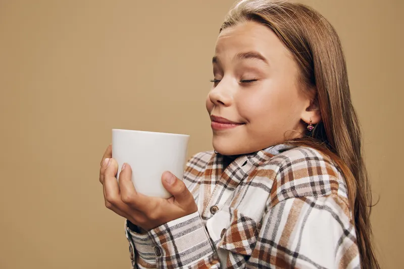 Girl holding white cup with both hands close to face enjoying warm drink with closed eyes wearing plaid shirt on beige background. Relaxation, comfort, beverage and casual look