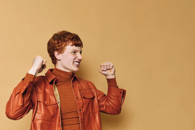Young redhead woman celebrating success with fists raised and happy face against beige background. Casual style with leather jacket and turtleneck in bright warm tones.