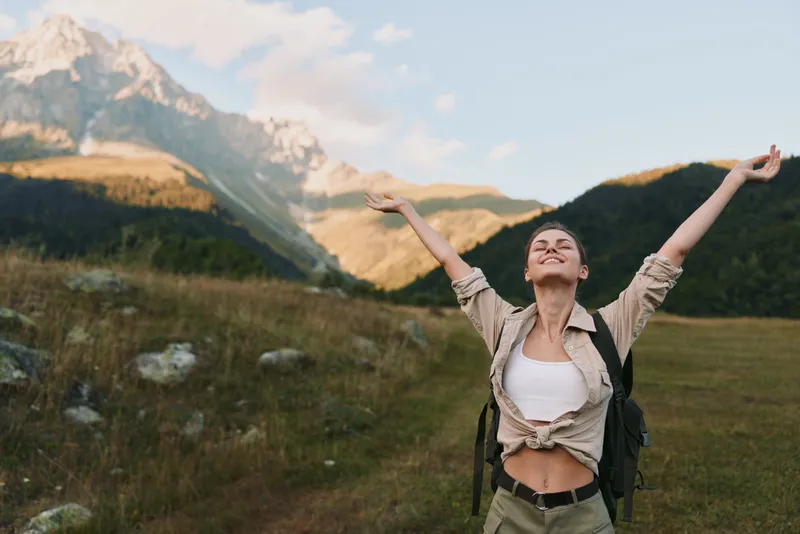 Woman traveler with backpack raises arms in triumph in a meadow beneath mountains, enjoying fresh air and sunlight, embracing nature and outdoor adventure in a carefree lifestyle