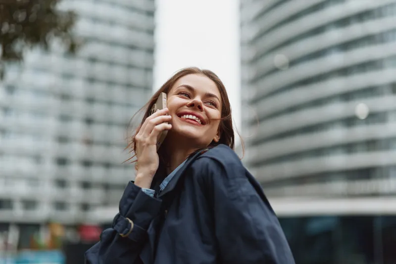 Smiling woman talking on smartphone outdoors in city. She wears dark coat and looks happy. Modern urban background with glass buildings and soft daylight.