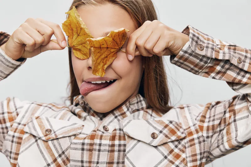 Child with autumn leaf covering eyes and tongue out, wearing plaid shirt with long hair in playful mood on white background. Cute kid enjoying fall season with fun expression.