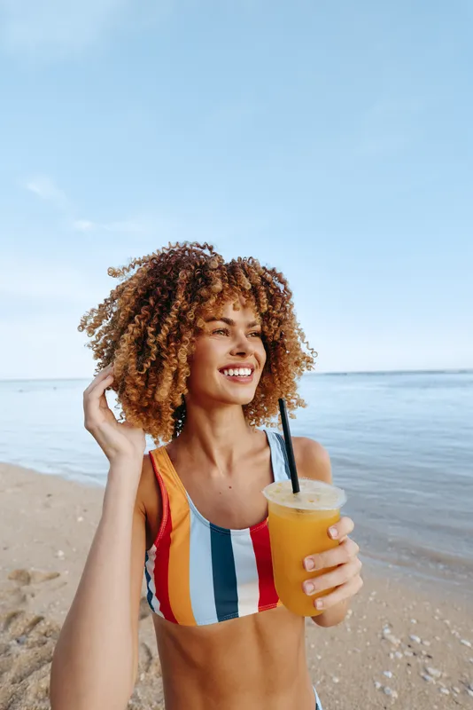 A smiling young woman with curly hair in a colorful striped bikini holds a cold orange drink on a sandy beach with calm sea and clear blue sky in the background.