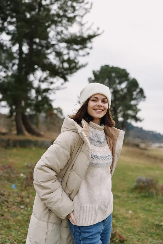 Winter outdoor portrait of a smiling woman in a warm beige coat, knitted sweater and beanie, standing in a grassy field with trees.