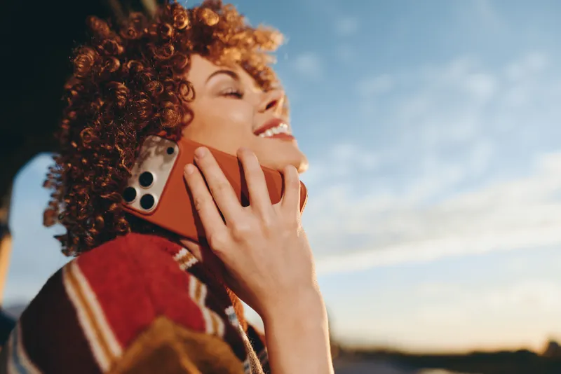 Woman with curly hair shows smile while holding smartphone outdoors, warm lifestyle moment, close up portrait, relaxed communication call, natural sunset light