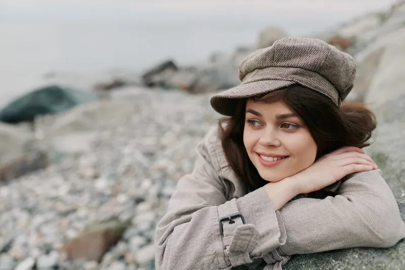 Young woman in casual clothing and hat smiles softly while resting on a rock at a pebble beach. Natural light creates a calm and peaceful mood in the outdoor setting.