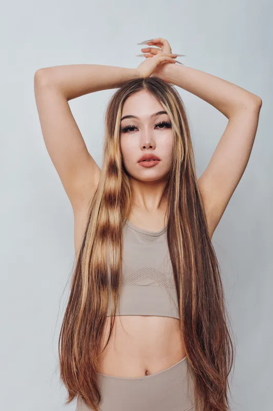 Woman with long straight hair poses with arms raised against plain background wearing sleeveless top. Beauty, fashion, hairstyle and confident look for portrait and style concept.