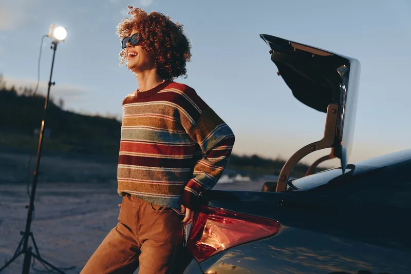Girl with curly hair by open car trunk outdoors at sunset, warm lifestyle portrait, casual striped sweater, golden backlight soft wind, filming light stand nearby, family travel atmosphere