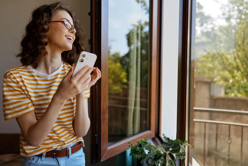 Young woman with curly hair in a yellow striped tee stands by an open window, smiling at her smartphone, capturing a cheerful, casual home moment.