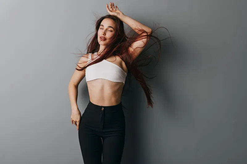 Young woman dancing with eyes closed and long hair flowing wearing white crop top and black pants against gray background in studio. Movement, freedom and style expression.