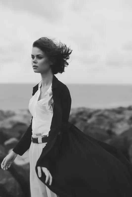 Woman walks along a windy coast in a flowing dress and blazer, sea in the background, dramatic monochrome mood, movement and outdoor fashion.