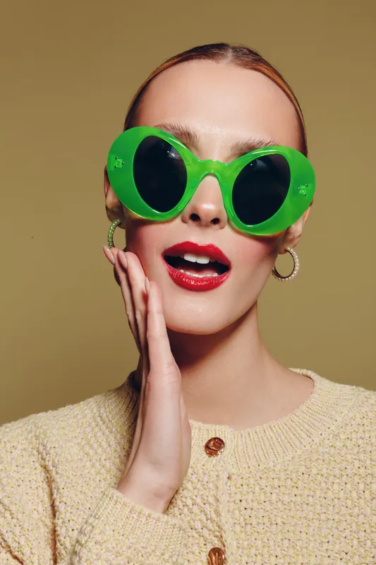 Young woman with green oversized sunglasses and red lipstick poses with hand near face on beige background. Fashion concept with bold accessories and stylish makeup.