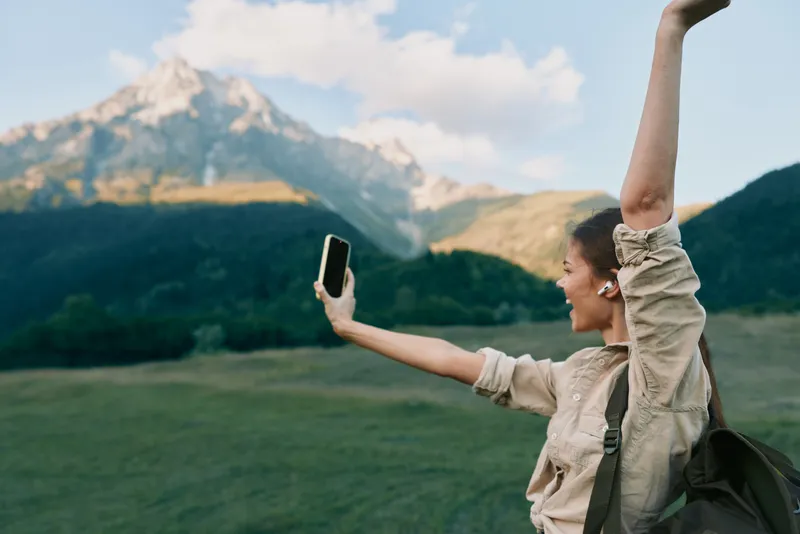 Young woman takes a selfie in a sunlit alpine meadow with towering mountains behind, expressing carefree happiness and a sense of freedom.