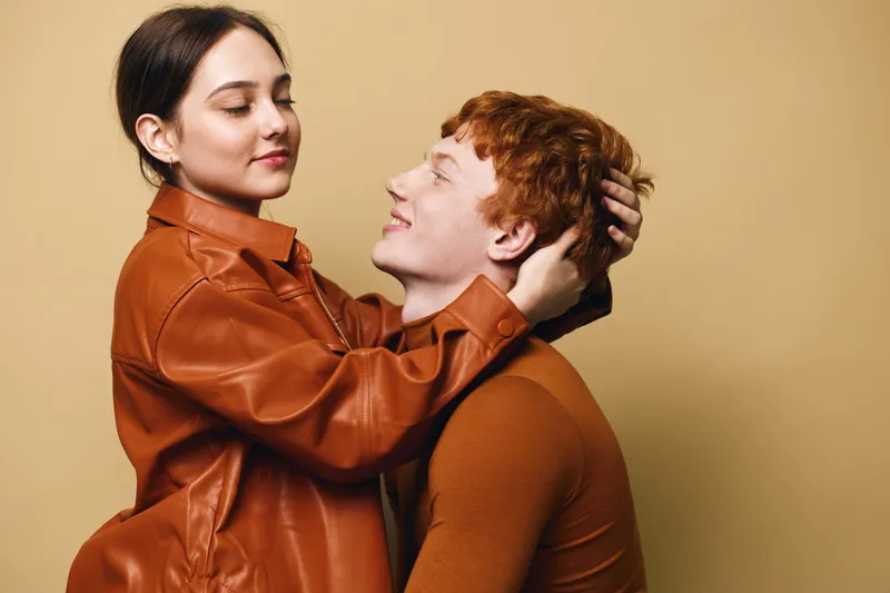 Young couple dressed in brown clothing embracing with affection in studio. Woman gently holds man face with love and connection in warm tone background.