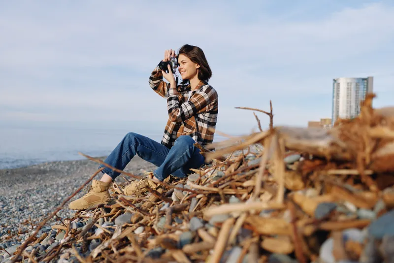 Photographer, outdoor scene with a woman using a camera on a rocky beach, driftwood, blue sky, natural light, candid moment, travel photography, serene mood, coastal landscape