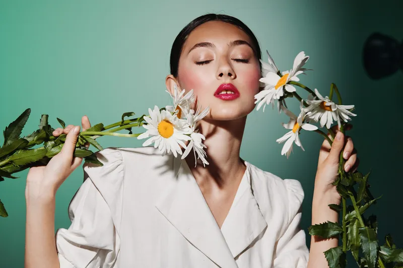 Young woman in white fashion coat holds large daisies with eyes closed, lips painted bright pink presenting beauty creative concept. Soft studio lighting with green background mood.