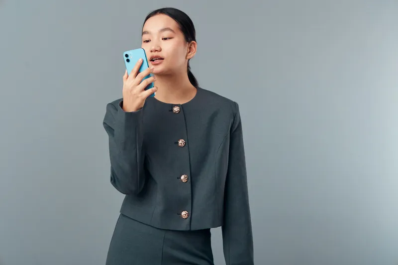 Young woman with smartphone speaking or recording voice message in studio. Female person using mobile phone with casual confident expression and plain background for technology communication.