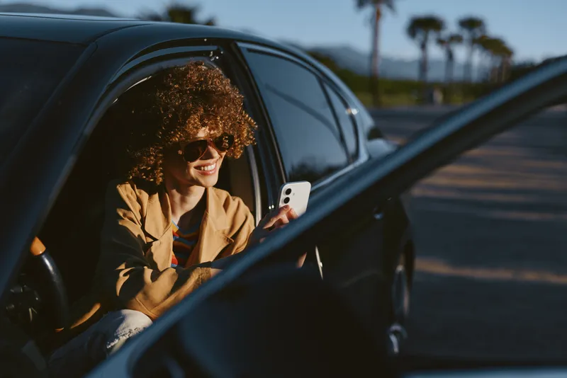A woman with curly hair sits in a car, looking at a smartphone, wearing a tan jacket. Bright outdoor light and a retro film color vibe emphasize casual, everyday mobility.