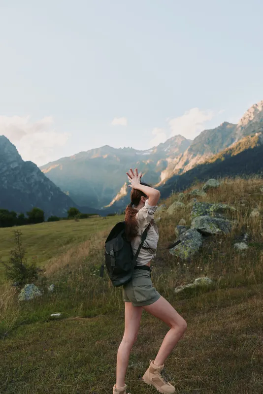 Young woman with long hair wearing hiking boots and a backpack enjoys a sunny mountain landscape while walking on a grassy trail during a casual outdoor adventure.