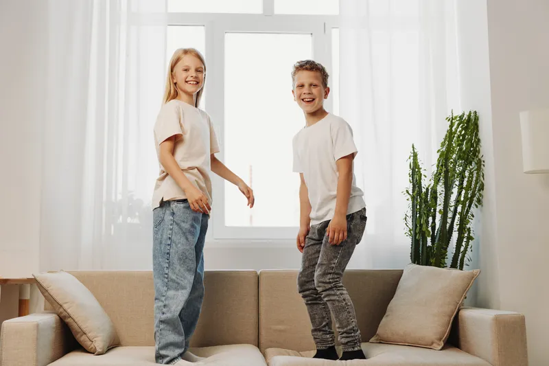 Two happy children jump on a beige sofa in a bright living room with large windows and green plants. The boy and girl wear casual clothes and share joyful moments together.