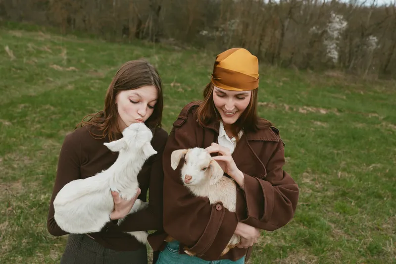 Two women in brown jackets cradle white goats in a green field, smiling and calm. A natural rustic lifestyle moment capturing gentle touch, companionship, and care for animals in a sunny countryside