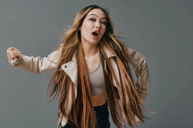 Young woman with long hair wearing casual jacket and gray top dancing and expressing joy on gray background in studio. Movement, fashion and happiness in portrait.