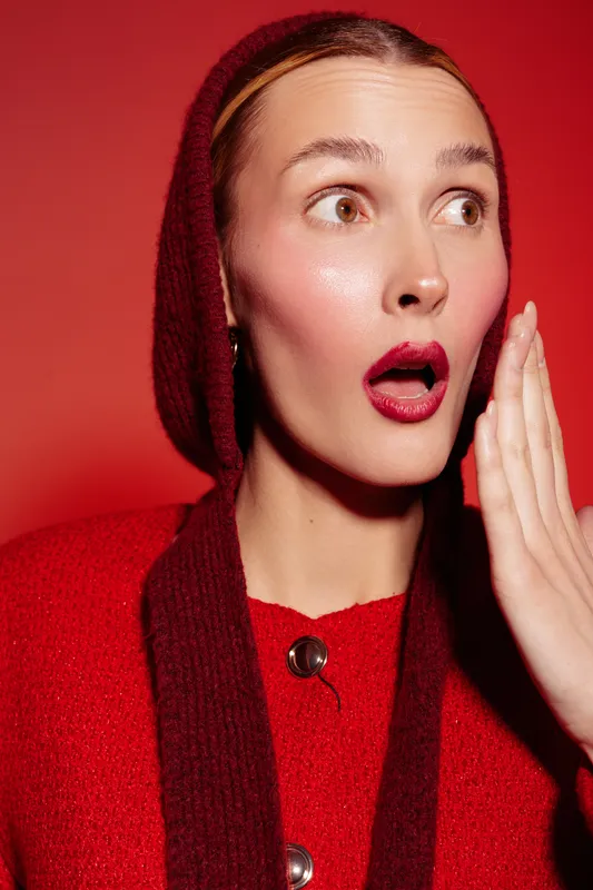 Surprised woman in red sweater and hood looks shocked with open mouth and hand near face on red background. Concept of emotion, reaction, and expression in portrait photography.