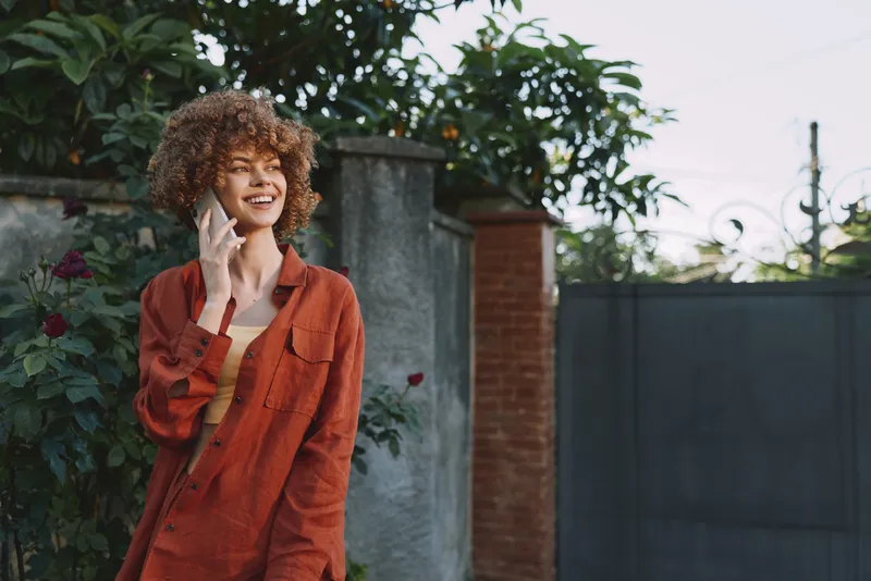 Smiling woman with curly hair in an orange jacket outdoors, talking on a phone. Natural daylight, casual lifestyle scene in a residential setting, warm, candid, everyday style and upbeat mood