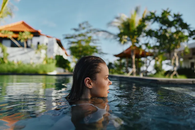 Relaxed woman enjoys a sunny poolside moment, gliding through clear water with a tropical resort backdrop, conveying leisure, calm, and vacation lifestyle in sunlight