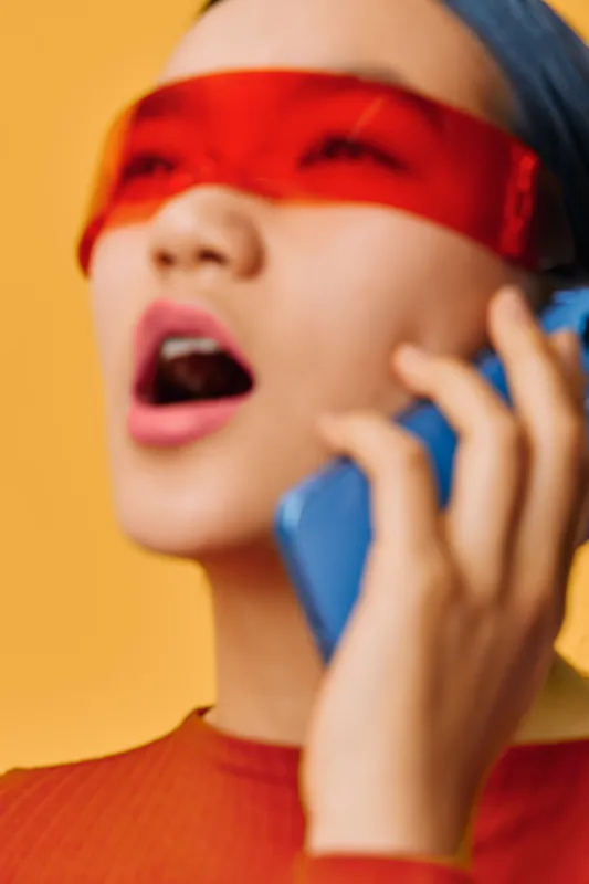 Woman with red visor and blue headband talking on smartphone with open mouth against orange background. Closeup portrait with blurred face and stylish modern accessories in vibrant colors.