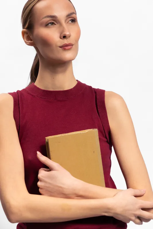 Young woman in sleeveless red top holds a closed book against her chest, looking thoughtfully to the side. Studio portrait with white background, concept of education and contemplation.