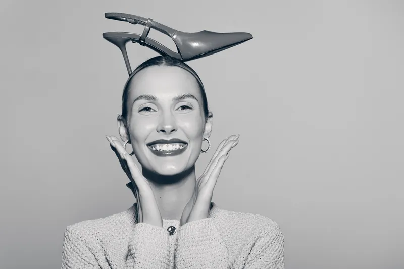 Happy Caucasian woman with light skin laughing and posing with a high heel shoe on her head in studio. Concept of fun, humor, and playful fashion accessory.