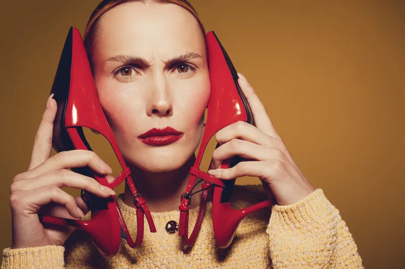 Woman with serious expression holds red high heels near face against brown background. Fashion concept with bold makeup and stylish shoes in studio lighting.