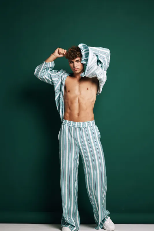 Young man with curly hair wearing striped pajamas posing with shirt open showing toned abs against green background. Casual fashion, relaxed style and confident expression.