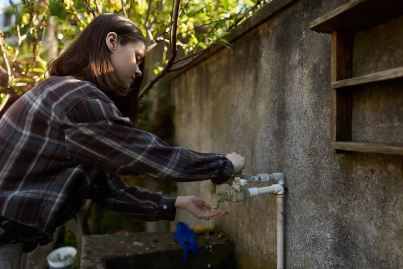 A woman in a plaid shirt performs an outdoor faucet task, capturing a casual, everyday moment. Natural daylight, wall backdrop, focus on hands and water flow