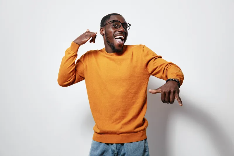African American man with dark skin wearing orange sweater and glasses smiling and pointing fingers on white background. Casual style and happy expression.