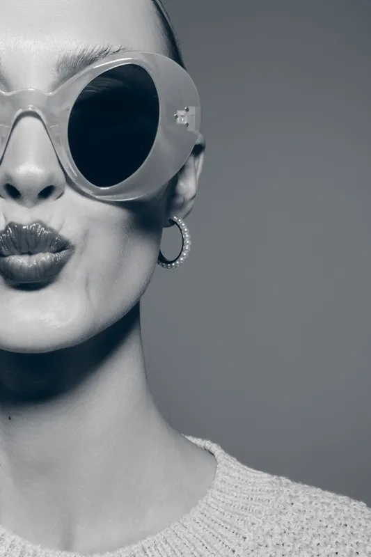 Young white woman with fair skin wearing large sunglasses and hoop earrings makes a playful kiss face. Close-up portrait in black and white with fashionable and stylish vibe.