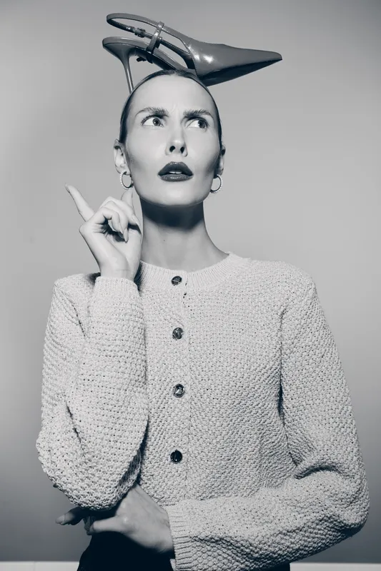 A fair skinned Caucasian woman wears a high heel shoe on her head, raising a finger while looking upward in a black and white studio portrait.