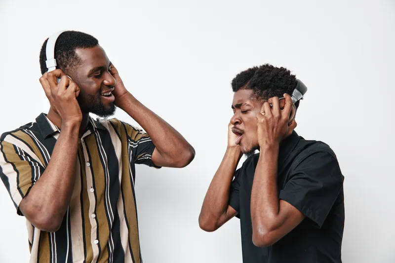 Two young black men with dark skin wearing headphones enjoying music together, smiling and interacting in casual clothing against white background in studio.