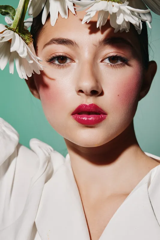 Close-up of a young woman with creative flower headpiece and vivid red lipstick. Smooth skin and fresh makeup emphasize beauty and fashion novelty.