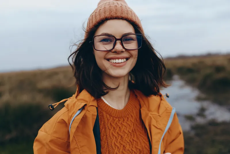 Smiling woman wearing orange knit sweater, glasses, brown coat and warm hat outdoors, walking on a rural path, lifestyle portrait in fresh autumn air near hills under blue sky today