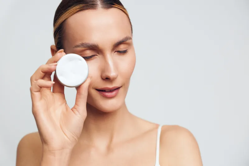Young woman with clear skin holding a white cotton pad near her face, eyes closed, demonstrating skincare routine, beauty concept, soft natural lighting on a white background.