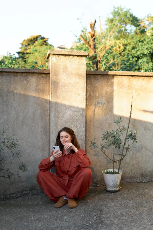 Young woman in a red jumpsuit sits on the ground beside a weathered wall, scrolling on her phone with a relaxed, thoughtful expression in warm sunlight