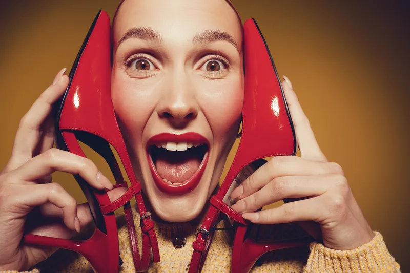 Excited woman with red lipstick holds red high heel shoes near face, smiling widely. Close-up portrait on yellow background. Concept of fashion, style, and joyful expression.