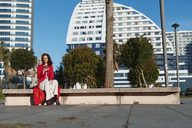 Young woman with curly hair wearing a red jacket and white pants sits on a concrete bench outdoors on a sunny day in an urban park setting.