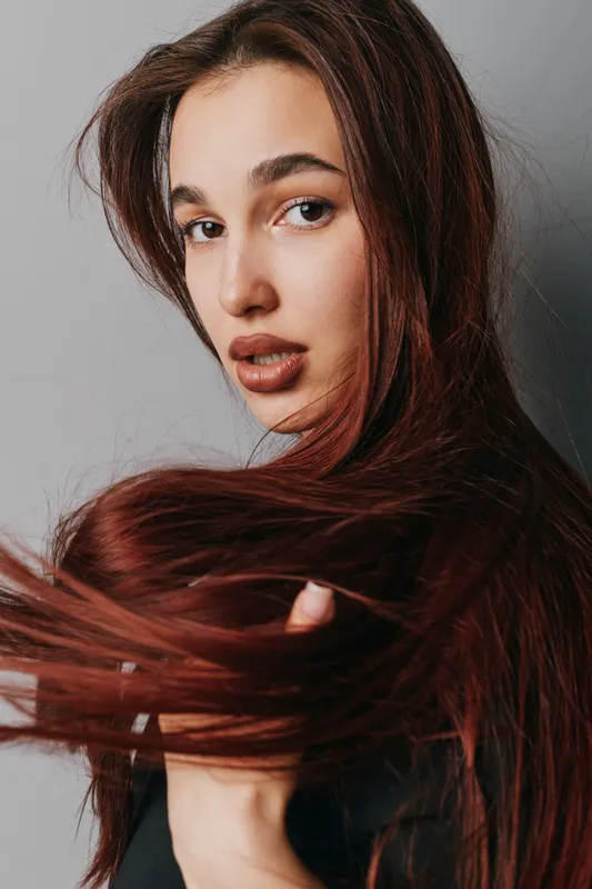 Woman with long brown hair looking over shoulder with serious expression against gray background. Female portrait with natural makeup and flowing hair in studio lighting closeup.
