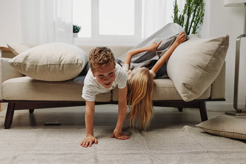 Playful children having fun on a beige sofa in a bright living room with natural light and cozy cushions. Concept of childhood joy, family moments, and home comfort.