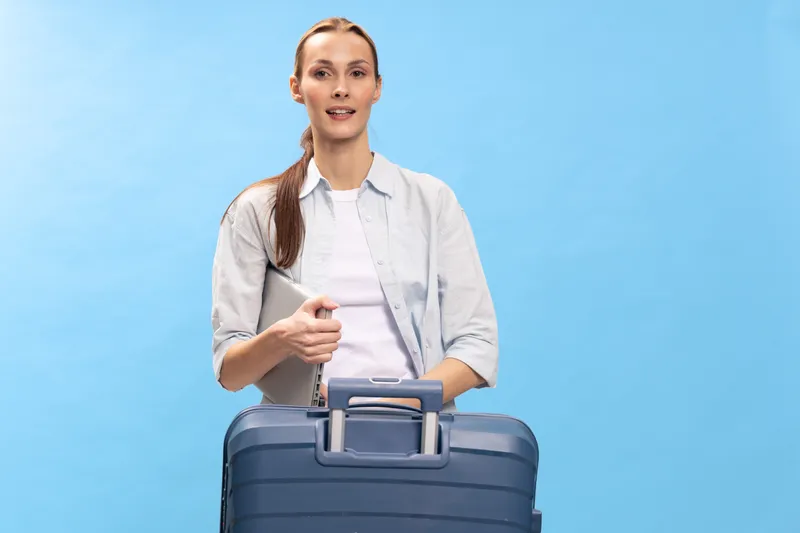 Young woman traveler with suitcase and laptop stands against blue background. Casual outfit and relaxed expression. Concept for travel, business trip, and lifestyle.