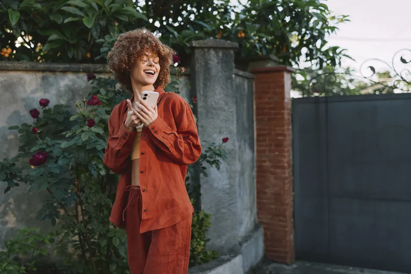 Curly haired woman outdoors wears a rust colored suit and laughs while checking her phone, standing beside a garden wall with blooming roses and soft afternoon light