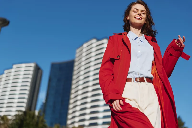 A stylish woman in a red coat walks confidently through a sunny urban street, creating a bold fashion moment for a contemporary city shoot.