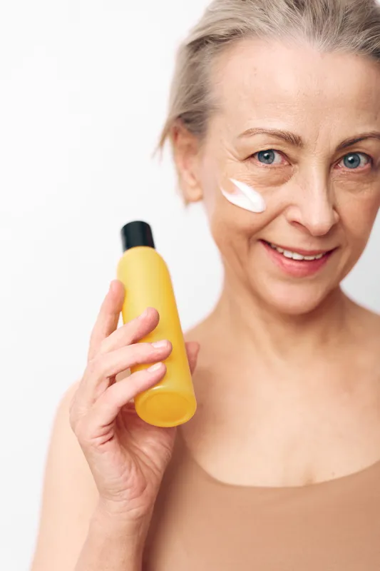Mature woman with blue eyes applies white face cream on cheek holding yellow bottle, smiling on light background. Concept of skincare, antiaging, and daily facial routine for healthy skin.